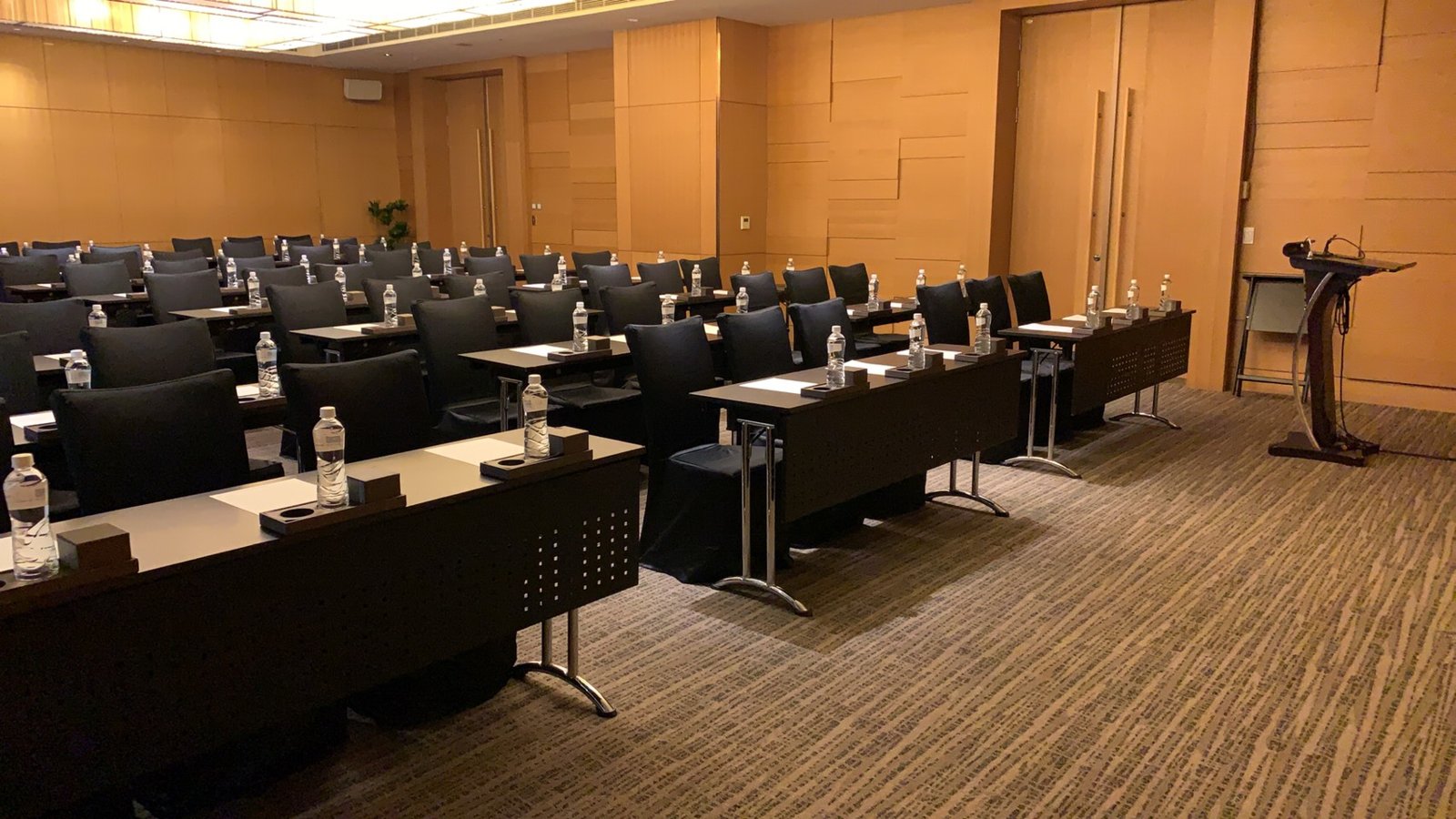 Rows of tables with water bottles in meeting room VEE05, with a lectern on the right